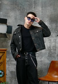 Jeune homme en veste et pantalon en jean foncé avec une chaîne, portant des lunettes de soleil, posant avec une main sur le front contre un mur gris texturé.