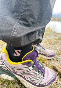 Violeta un melna sporta apava ar dzelteniem akcentiem, ar tīkliņa augšdaļu un šņorējuma sistēmu. Melna zeķe ar rozā logotipu. Redzama fakturēta auduma un detaļas.