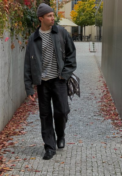 Hombre con chaqueta negra, camisa a rayas y gorro caminando por un camino de adoquines, rodeado de hojas de otoño y vegetación.