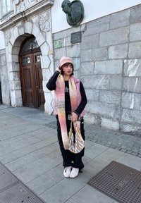 Jeune femme portant un gilet rose, un bonnet en maille et une longue écharpe, debout sur un trottoir en pierre près d'une porte en bois ornée d'arcs et d'un mur en pierre grise avec une plaque.