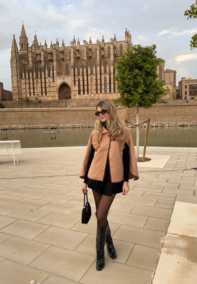 Mujer con vestido negro y capa beige sosteniendo un bolso cerca de un canal, con una gran catedral gótica de piedra en el fondo.