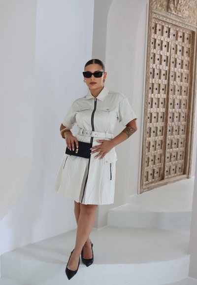 Mujer con el cabello peinado hacia atrás, vistiendo un vestido blanco ceñido con cinturón, gafas de sol negras y tacones, sosteniendo un bolso negro en un interior blanco minimalista.