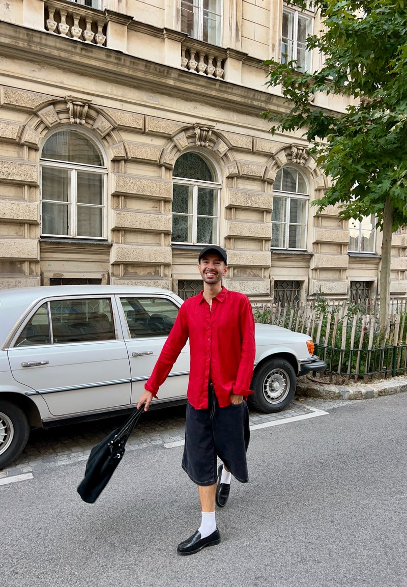 Camisa vermelha de botões, saia preta, meias brancas, sapatos pretos e uma bolsa preta. Passeio perto de um carro vintage, edifício bege com janelas em arco.