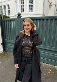 Abrigo negro con solapas anchas sobre un top negro de encaje, combinado con pantalones negros de talle alto, posando frente a un fondo de cerca de madera verde.