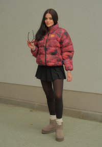 Πουπουλένιο μπουφάν σε κόκκινο και μαύρο tie-dye σχέδιο, με φερμουάρ, σε συνδυασμό με μια μαύρη φούστα, μαύρες καλτσοδέτες και γκρίζες πλατφόρμες με λευκές κάλτσες.