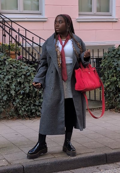Abrigo de lana gris, blusa blanca, corbata burdeos, falda gris, leggings negros, botas de plataforma negras y un bolso rojo contra un fondo de follaje verde.