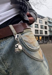 Άτομο που φοράει φθαρμένα τζιν, με ασορτί αλυσίδα και ένα κρεμαστό κόσμημα σε σχήμα καρδιάς προσαρτημένο σε καφέ ζώνη, στέκεται έξω κοντά σε κτίρια.