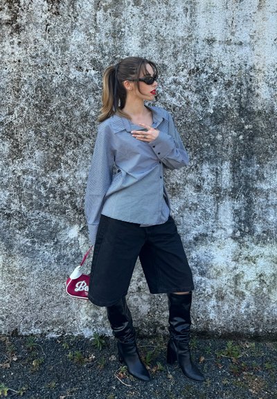 Mujer con coleta, gafas de sol, camiseta a rayas azul, pantalones cortos negros hasta la rodilla, botas de tacón negras y bolso rojo de pie frente a una pared texturizada.