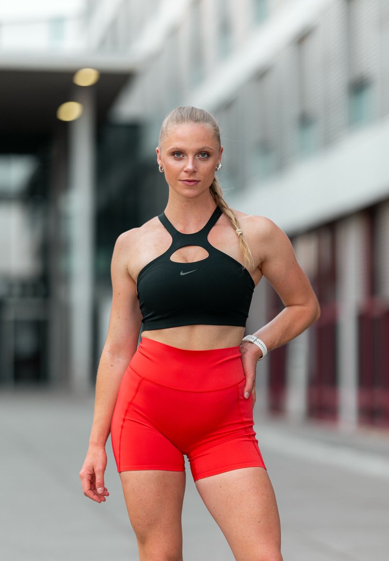 Trænet kvinde med fletninger iført en sort sportstop og røde shorts står udendørs med den ene hånd på hoften og ser på kameraet.