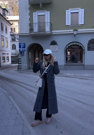 Mujer vestida con un abrigo gris largo, gafas de sol, gorra blanca y botas beige se encuentra en una calle curva cerca de edificios históricos con ventanas cerradas.