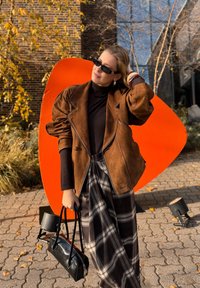 Hnedá semišová nadrozmerná bunda s kontrastným čiernym prešívaním, čierny rolák, kárované široké nohavice a čierna kabelka; oranžové pozadie.