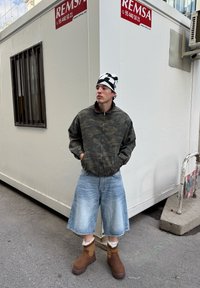 Vyras dėvintis maskavimo striukę, plačias džinsines šortus, rudus batus ir juodai baltą kepurę, stovintis šalia balto statybos konteinerio gatvėje.