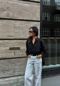 Sort knop-bluse, hvide vide bukser, guld accessories, solbriller, står foran en struktureret stenvæg med en reflekterende glasoverflade.