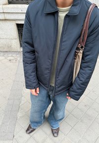 Doudoune bleu marine avec fermeture éclair et poches latérales, portée sur une chemise vert clair. Associée à un jean bleu ample et des chaussures marron.