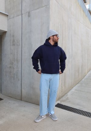 Hombre con gorro gris, sudadera con capucha azul marino, jeans azul claro y zapatos tipo slip-on a cuadros, de pie sobre concreto cerca de una esquina de pared.