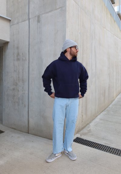 Hombre con gorro gris, sudadera con capucha azul marino, jeans azul claro y zapatos tipo slip-on a cuadros, de pie sobre concreto cerca de una esquina de pared.