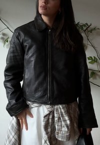 Giacca di pelle nera con colletto, chiusura con zip e maniche lunghe. Indossata sopra un vestito bianco con una cravatta a quadri in vita.