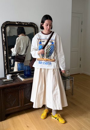 Mujer en el interior con falda blanca, camisa de manga larga con estampado de pájaros, zapatillas amarillas y bolso bandolera, de pie cerca de un mueble antiguo y un espejo.
