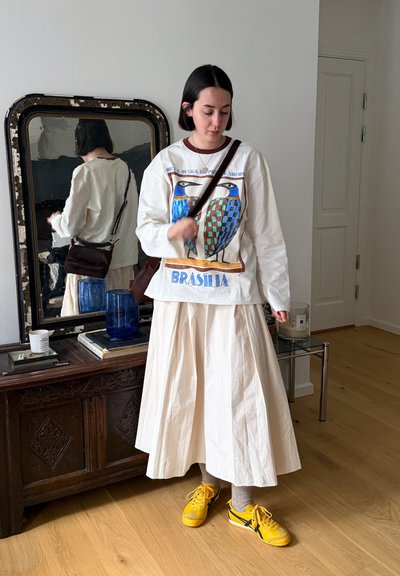 Mujer en el interior con falda blanca, camisa de manga larga con estampado de pájaros, zapatillas amarillas y bolso bandolera, de pie cerca de un mueble antiguo y un espejo.