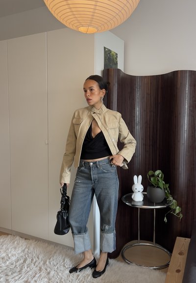 Mujer con cabello oscuro, chaqueta beige, top negro, vaqueros remangados y zapatos planos negros, de pie en el interior cerca de una mesa redonda con una lámpara de conejo blanco y una planta.