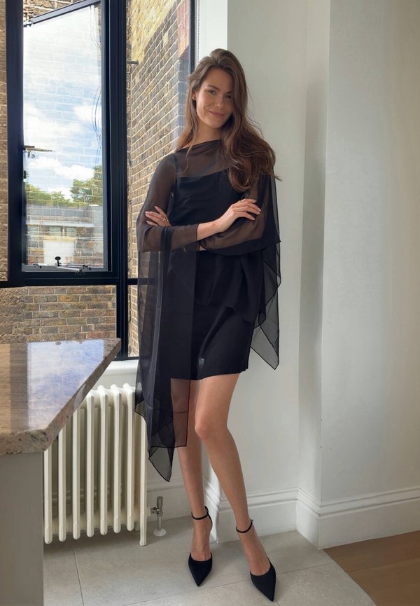 Mujer de pie en el interior cerca de una ventana, vistiendo un vestido negro transparente y tacones altos negros puntiagudos, sonriendo con los brazos cruzados.