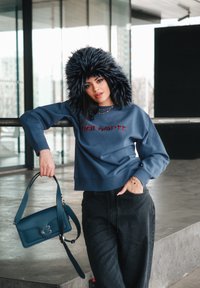 Bluet Sweatshirt mit rotem Logo, schwarze Fake-Fur-Mütze, dunkle Jeanshose und eine türkisfarbene Handtasche mit strukturiertem Design, präsentiert in einer städtischen Umgebung.