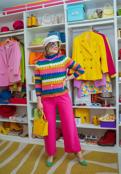 Mujer con suéter a rayas de arcoíris, pantalones rosas, tacones verdes, sombrero blanco, gafas de sol de colores, sosteniendo bolso amarillo, de pie en un armario colorido.