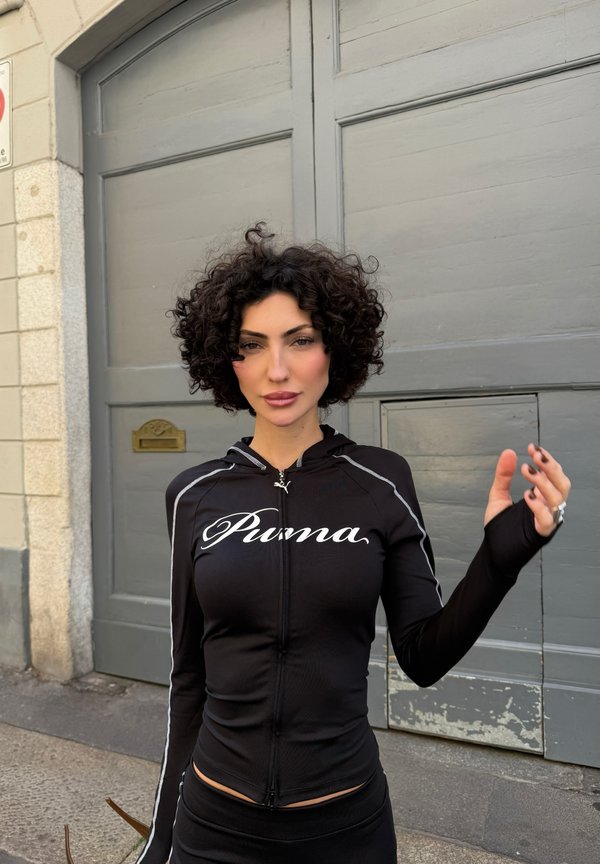 Femme aux cheveux bouclés portant une veste zippée noire Puma, debout devant une grande porte en bois grise, levant la main droite.