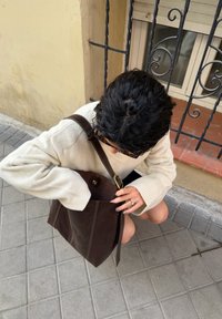 Personne aux cheveux foncés et portant un pull clair à genoux sur le trottoir, atteignant un grand sac à bandoulière en cuir brun foncé près d'un bâtiment avec une fenêtre grillagée.