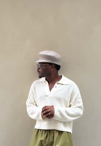 Hellbeiger Strickpullover mit einem Kragen und drei Knöpfen, kombiniert mit olivgrünen Hosen; trägt eine hellbeige Mütze und eine Brille.