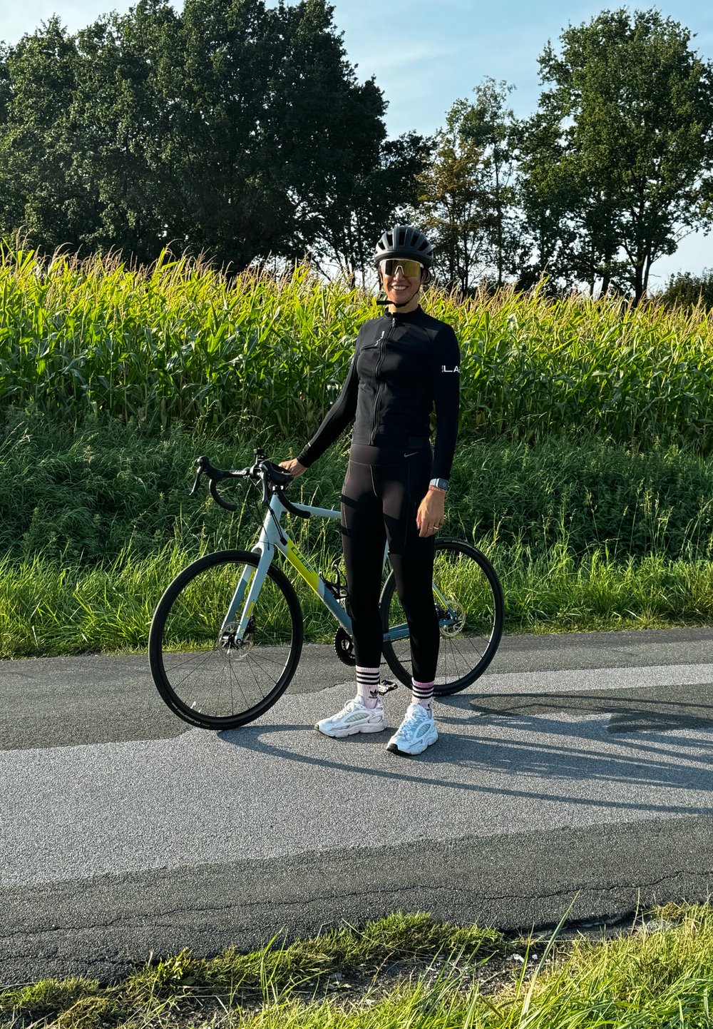 Racefiets met een grijs en groene frame. De fietser draagt een zwart fietsoutfit, witte sneakers en gestreepte sokken, staande op een verharde pad.