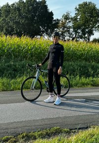 Racefiets met een grijs en groene frame. De fietser draagt een zwart fietsoutfit, witte sneakers en gestreepte sokken, staande op een verharde pad.