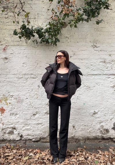 Mujer con chaqueta acolchada negra, camiseta negra de Adidas, pantalones negros y gafas de sol, de pie sobre un suelo cubierto de hojas frente a una pared blanca de ladrillo con hiedra.