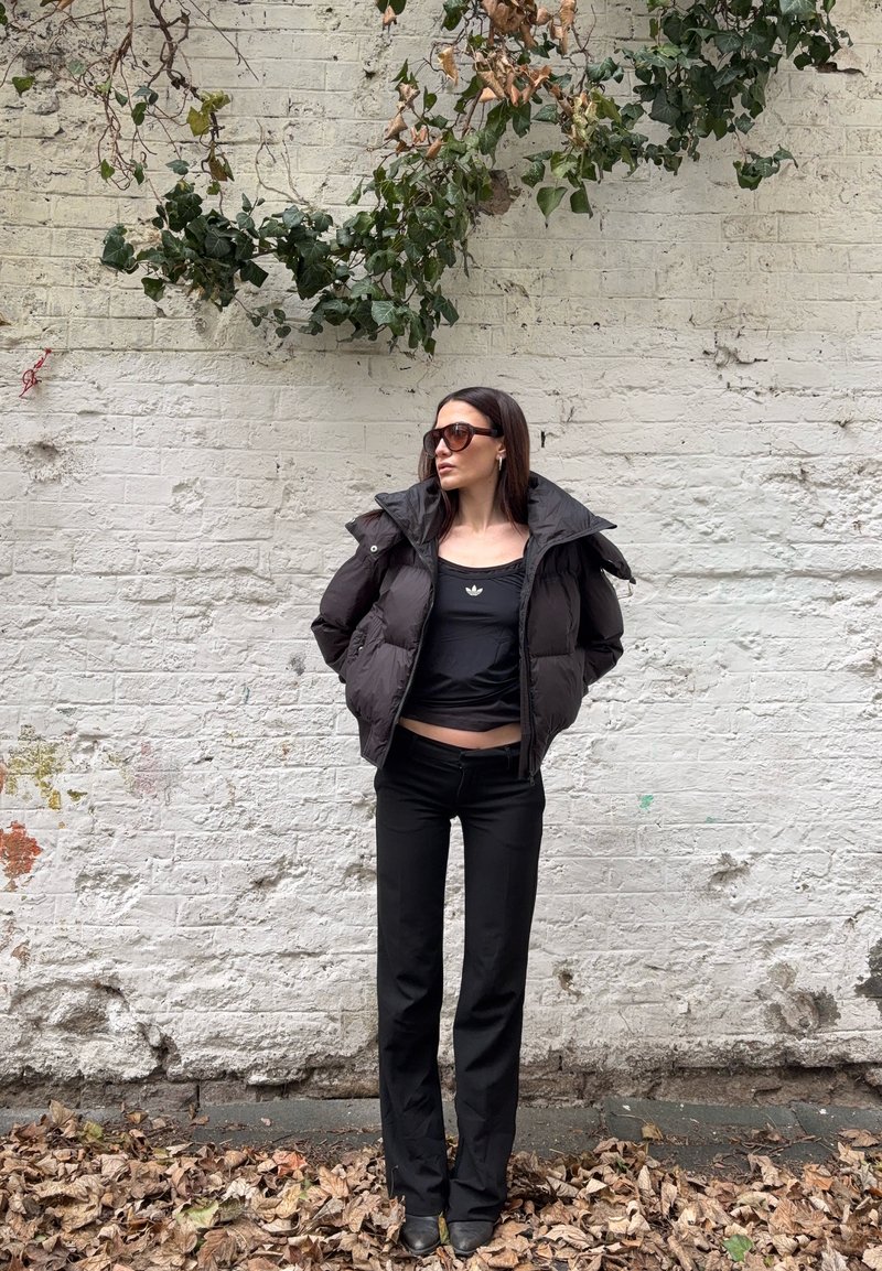 Žena odjevena u crnu pernatu jaknu, crni Adidas gornji dio, crne hlače i sunčane naočale stoji na tlu prekrivenom lišćem nasuprot bijelog zidnog zida s bršljanom.