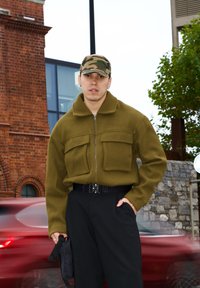 Chaqueta verde oliva con cremallera y dos bolsillos delanteros, pantalones anchos negros y una gorra de camuflaje, sosteniendo una pequeña bolsa negra. Entorno urbano con un coche rojo en movimiento.