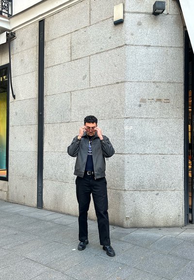 Chaqueta de cuero negra sobre una camisa oscura, pantalones negros y zapatos negros. De pie frente a una pared de piedra gris con el letrero 'ETRO'.