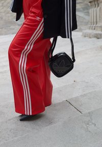 Pantalons larges rouges avec des rayures blanches sur les côtés, associés à un sac carré noir arborant un logo, le tout sur des marches en pierre. Des chaussures à talons noires sont portées.