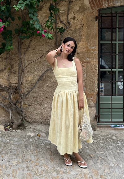 Vestido amarillo claro, hasta la rodilla, con textura acanalada y escote cuadrado. Combinado con chanclas blancas y un bolso brillante. Fondo de pared de piedra.