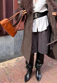Personne portant un manteau bordé de fourrure marron foncé, chemise blanche, large ceinture noire, jupe grise, bottes noires pointues, tenant un sac à main en cuir marron.