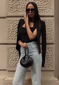 Črn prilegajoč se blazer preko črnega topa in svetlo modrih hlačah z visokim pasom. Model drži črno ukrivljeno torbico z gladko površino.