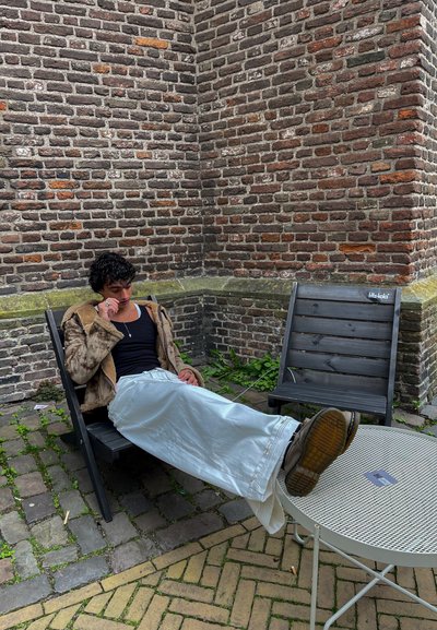 Persona relajada con una chaqueta forrada de piel color beige, top negro y pantalones blancos, sentada en sillas de madera negras junto a una mesa metálica redonda. Fondo de pared de ladrillo.