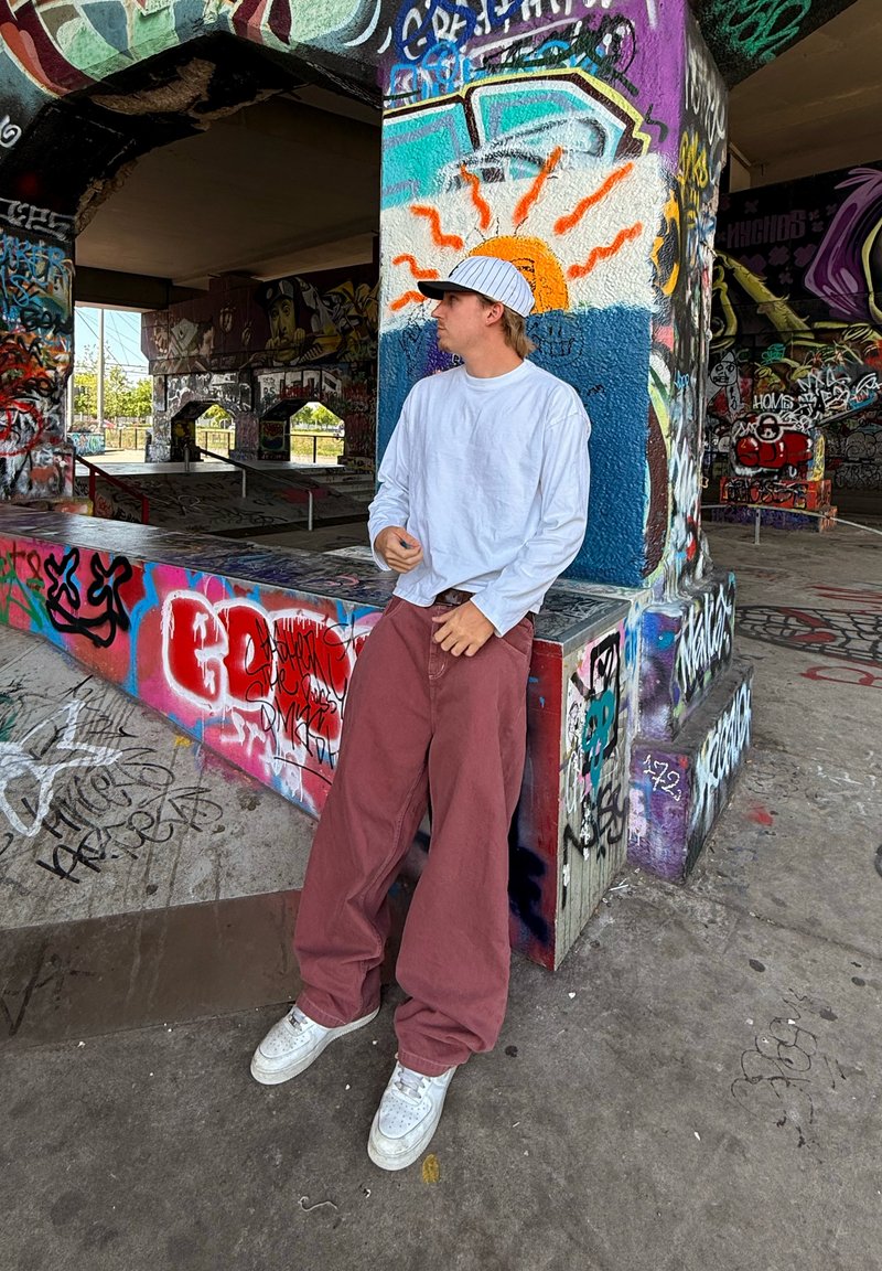 Hvid langærmet skjorte, maroon posede bukser og hvide sneakers. Står ved siden af en graffiti-dækket søjle i en skatepark.
