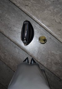 Μαύρη δερμάτινη τσάντα με χρυσό φερμουάρ, τοποθετημένη σε μια υφασμάτινη γκρι πέτρα δίπλα σε ένα διάφανο ποτήρι με χρυσή κυκλικ�ή βάση και κοφτερά μαύρα τακούνια.