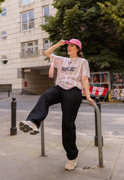 Camiseta Nike a rayas rosas y blancas, pantalones de mezclilla negros de gran tamaño, gorra rosa y zapatillas blancas con detalles grises. El escenario es una calle urbana.