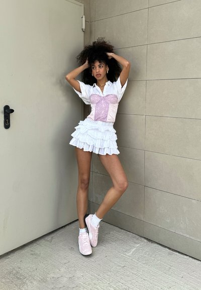 Camisa blanca con corsé rosa, falda de volantes blanca en capas y zapatillas rosas. Cabello rizado, de pie contra una pared lisa.