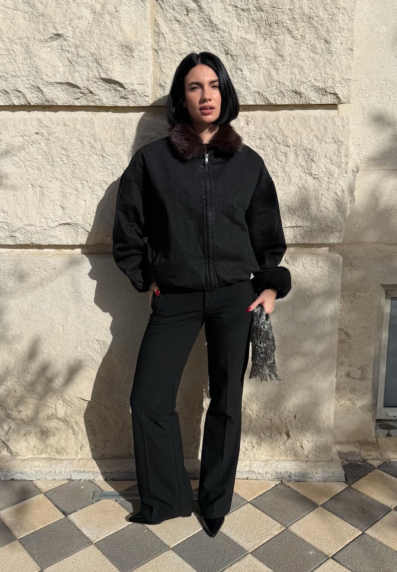 Femme avec une coupe au carré noire portant une veste noire à col en fourrure, un pantalon noir, des chaussures pointues, tenant une pochette argentée à perles, debout contre un mur en pierre.