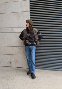 Oversized sort og brun camouflage sweater, beige vest, blå jeans og mørkebrune ankelstøvler, afsluttet med en grøn strikket hue.