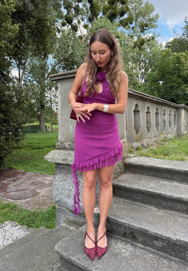 Donna con abito viola e scarpe abbinate che sta su gradini di pietra all'aperto, aggiustando degli anelli con alberi e ringhiera in pietra sullo sfondo.