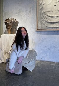 Beige håndveske med et enkelt design holdt av en person i en teksturert hvit genser og et skimrende sølvskjørt. Bakgrunnen har kunst og keramikk.