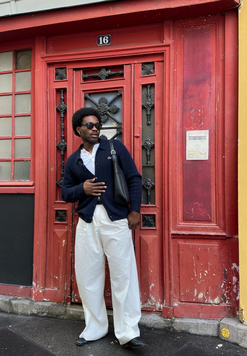 Hombre con gafas de sol, cárdigan azul marino, pantalones blancos y zapatos negros, de pie frente a una puerta roja ornamentada con el número de casa 16.